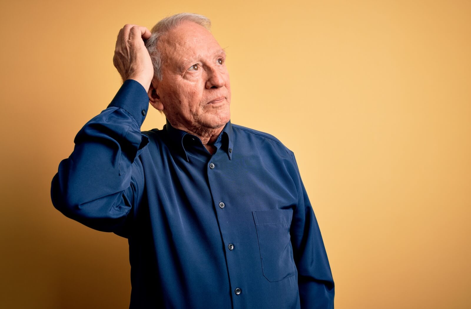 An older adult in a navy blue shirt scratching their head with a puzzled expression, appearing confused or uncertain.