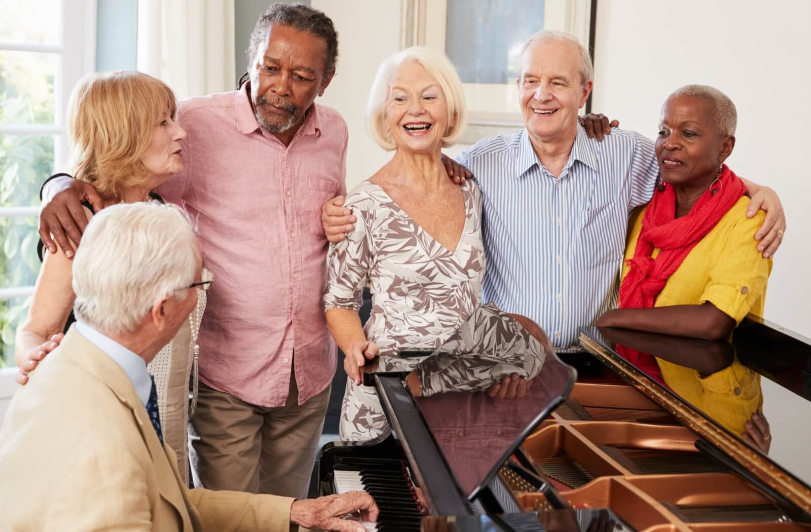 A group of older adults arm-in-arm around a piano in the common room of a senior living community, singing and smiling together.