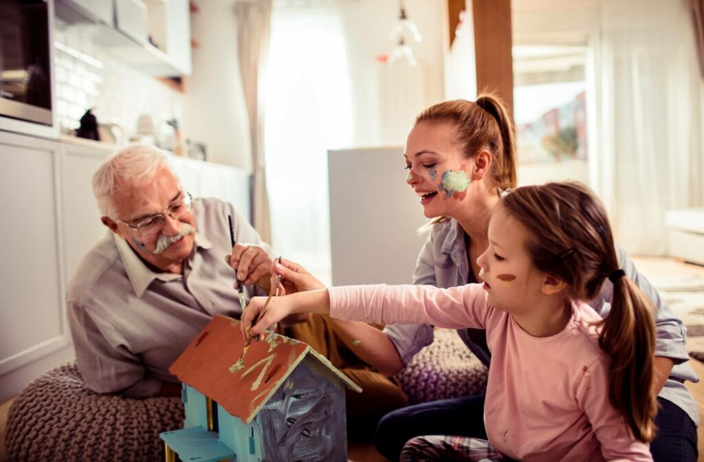 An older adult happily paints a doll house with their adult child and grandchild during a visit to their memory care home.