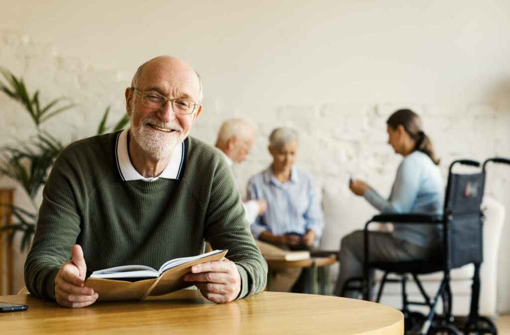 An older adult man sitting at a table in a common area looking up and smiling while holding a book. In the background, another group of seniors sits around a table, one in a wheelchair.